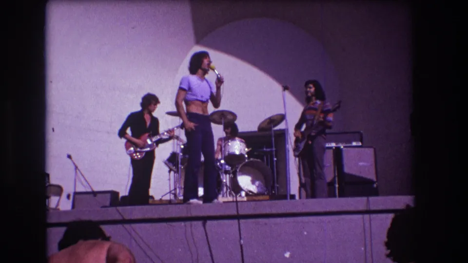 1973 Medford, New Jersey concert frame showing the singer at center with guitarist, bassist, drummer, microphones, and amplifiers around him.