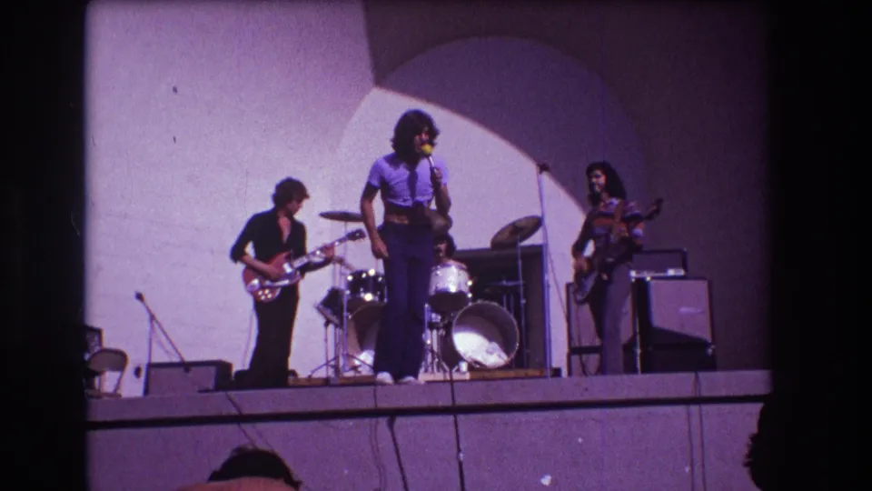 1973 Medford, New Jersey 8mm frame with the vocalist near center stage, a red electric guitar at left, drums behind, and bass at right.