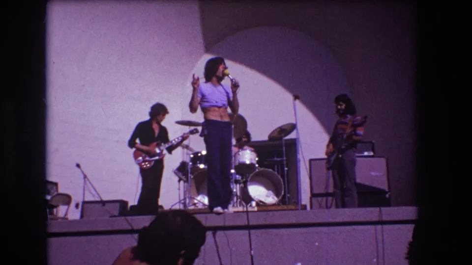 1973 Medford, New Jersey home-movie frame of the singer gesturing on an outdoor stage with guitarist, drummer, bassist, and amplifiers visible.