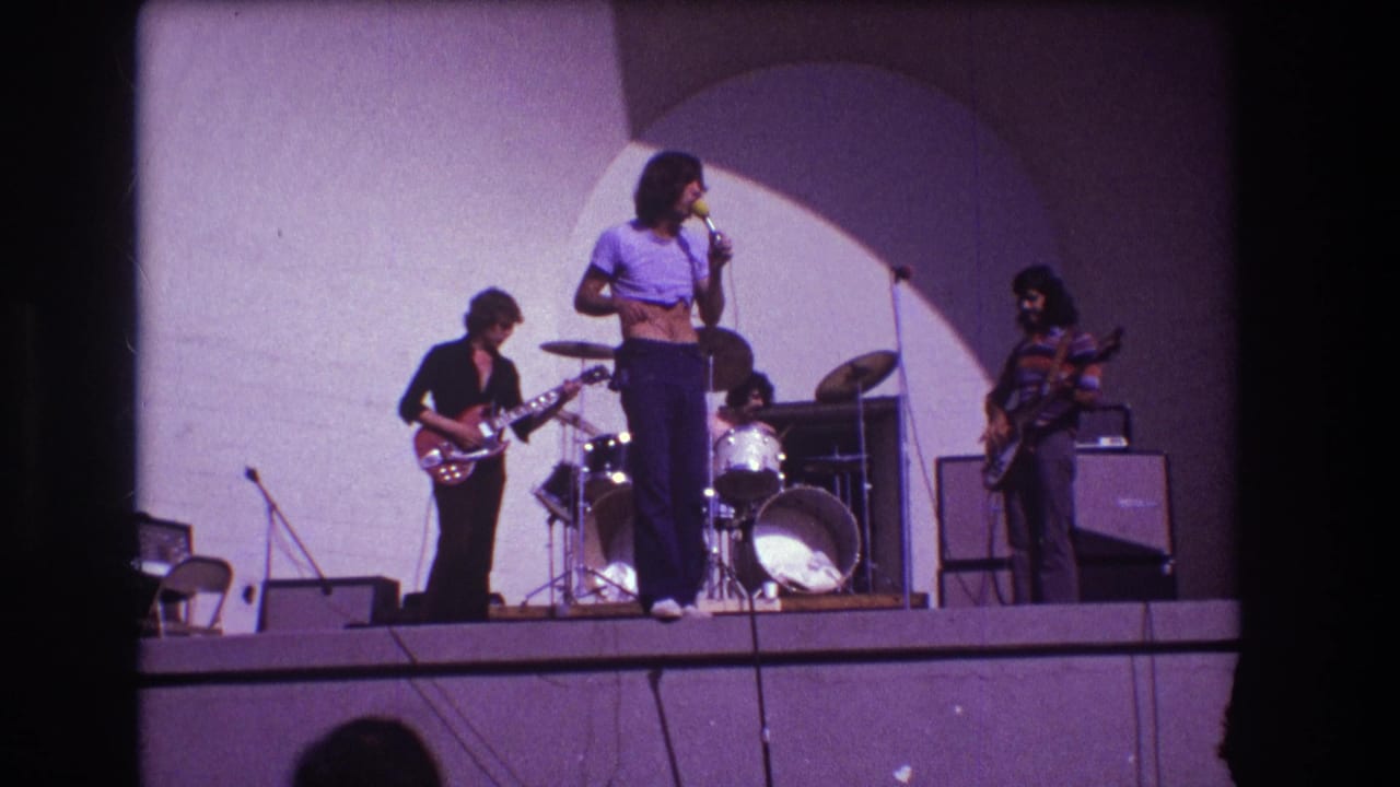 Rock band on a small stage with singer at microphone, guitarists, drummer, amplifiers, and spotlighted back wall.