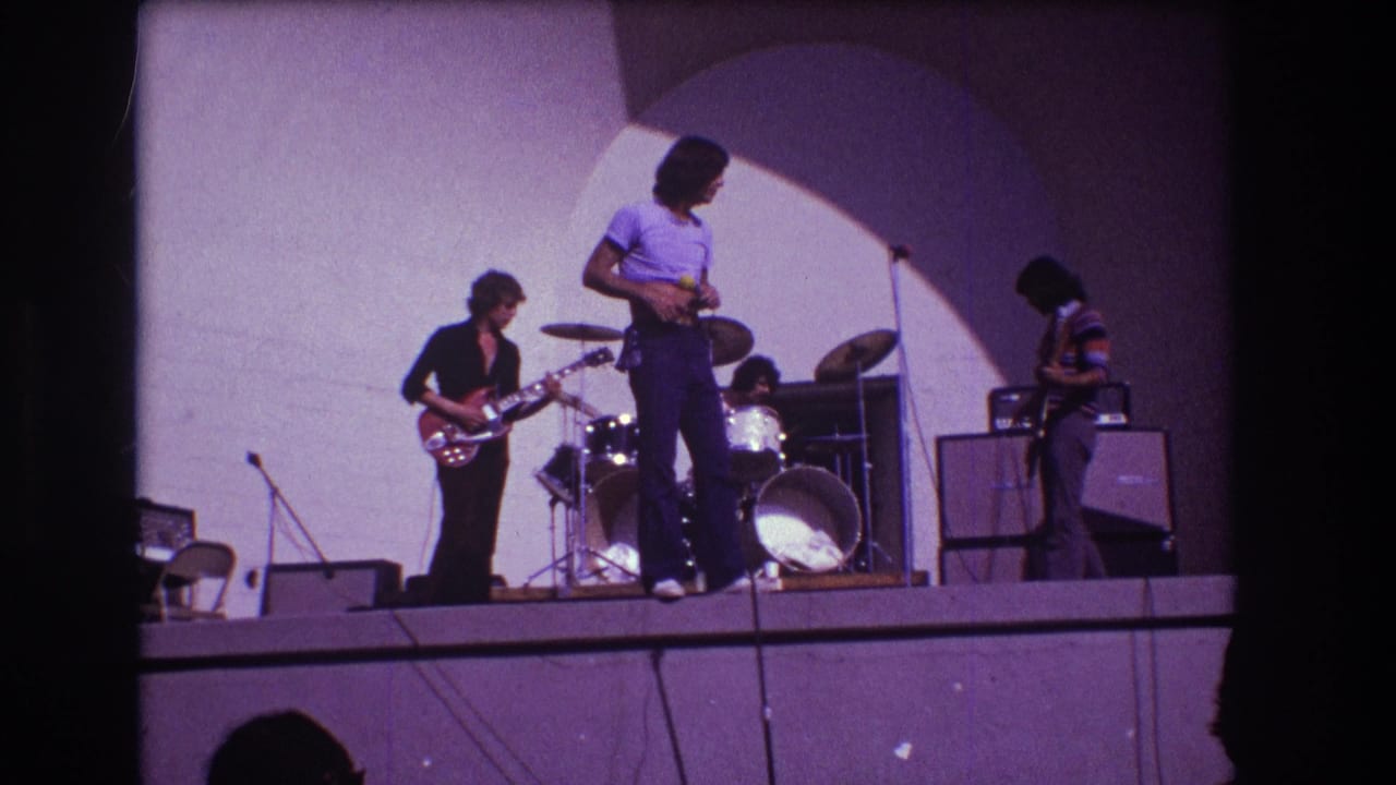 Singer turns on stage while guitarists and drummer continue playing beside amplifiers and a lit wall.