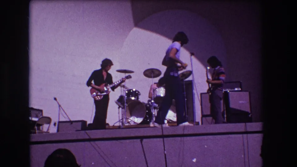 Home-movie frame from the 1973 Medford performance showing the singer moving across the stage between guitarists and the drum kit.
