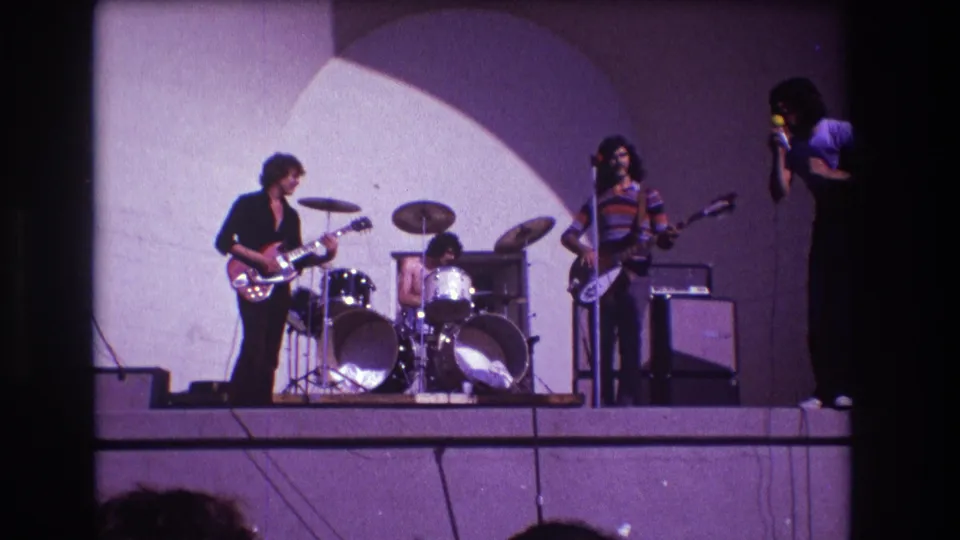 1973 Medford 8mm frame showing drummer and guitarists on a small stage, with the singer partly visible at the right edge holding a microphone.
