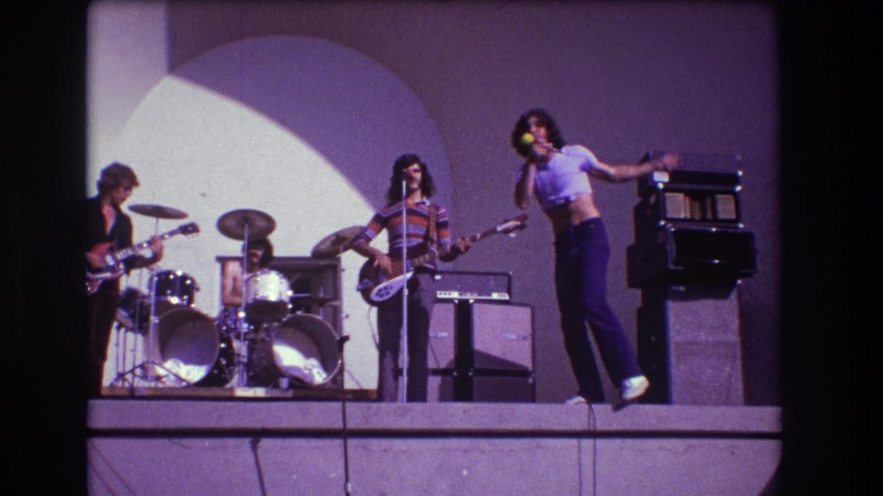 In 1973 Medford, New Jersey footage, a rock band performs on stage with a singer at right, guitarist at left, drummer center, and bassist near a microphone.