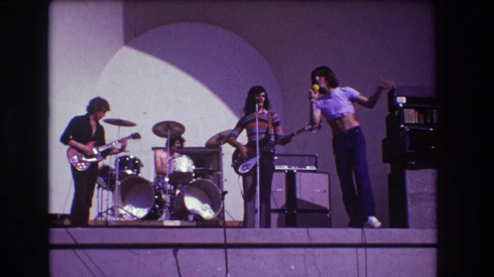 In the Medford, New Jersey home-movie footage, the singer gestures near the stage edge while the rest of the rock band performs under colored light.