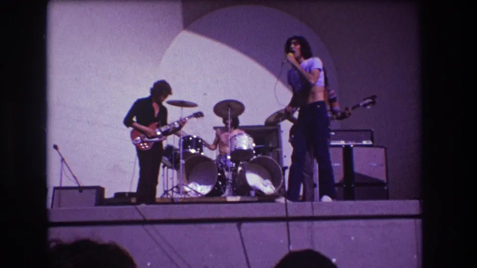 1973 Medford, New Jersey frame of a live band performance with singer, drummer and guitarist under an arched backdrop.