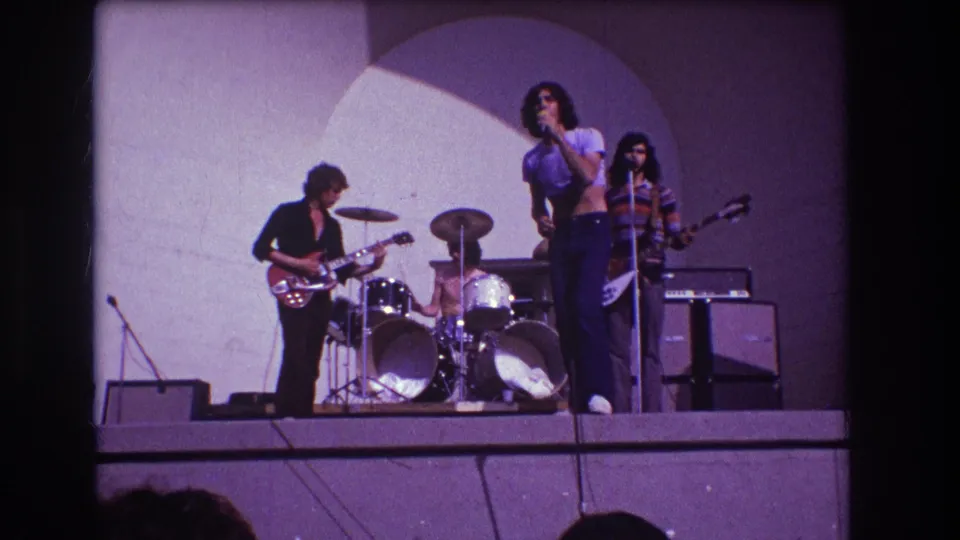 1973 Medford, New Jersey home-movie frame showing a rock band on stage with singer at microphone, guitarist, drummer, bassist, and amplifiers.