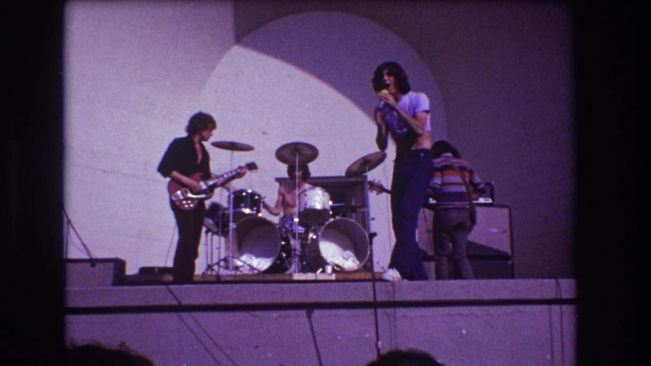 Rock band on an outdoor stage with guitarist, drummer, singer, and bassist near amplifiers.