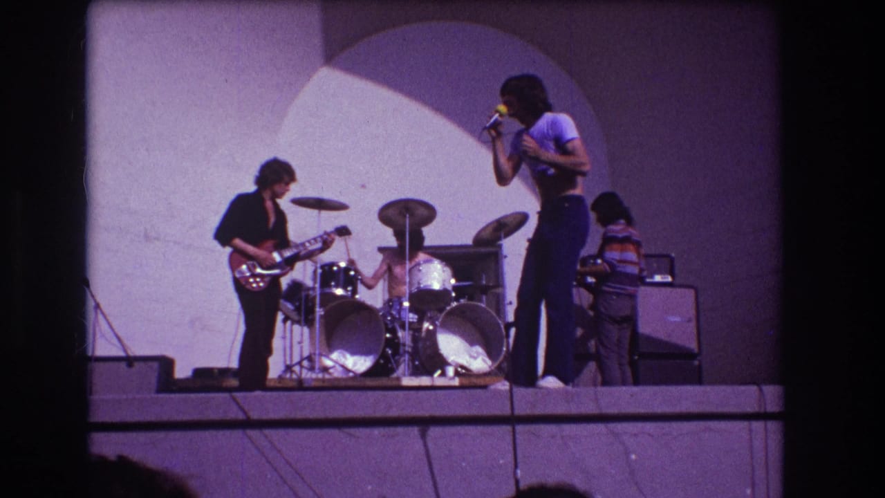 Singer holds a microphone while guitar, drums, bass, and amplifiers fill the stage.