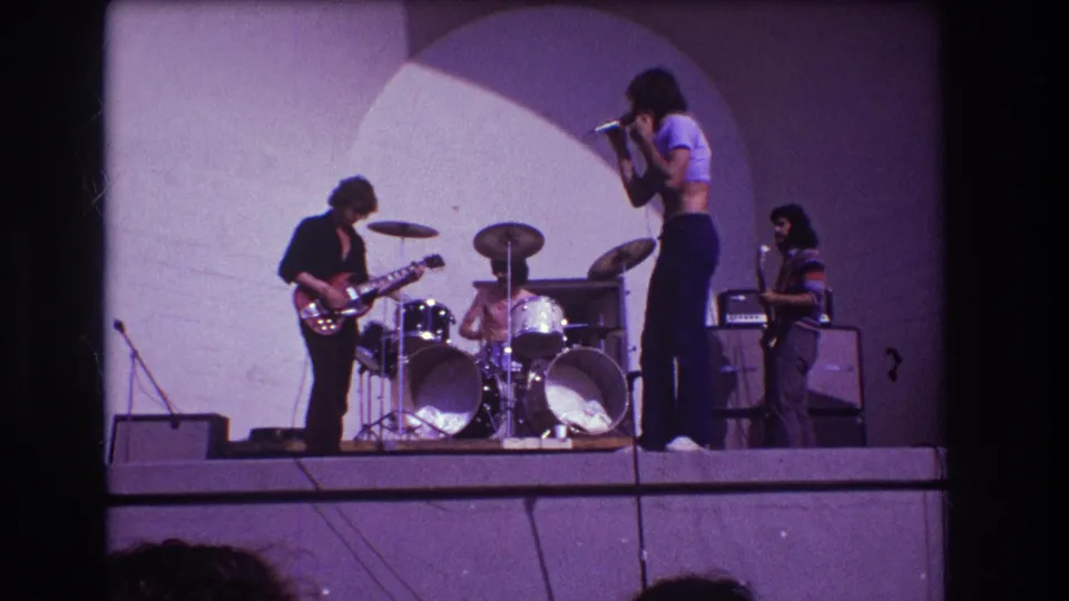 1973 Medford, New Jersey home-movie frame showing the singer in profile beside guitar, drums, bass, and stage equipment.