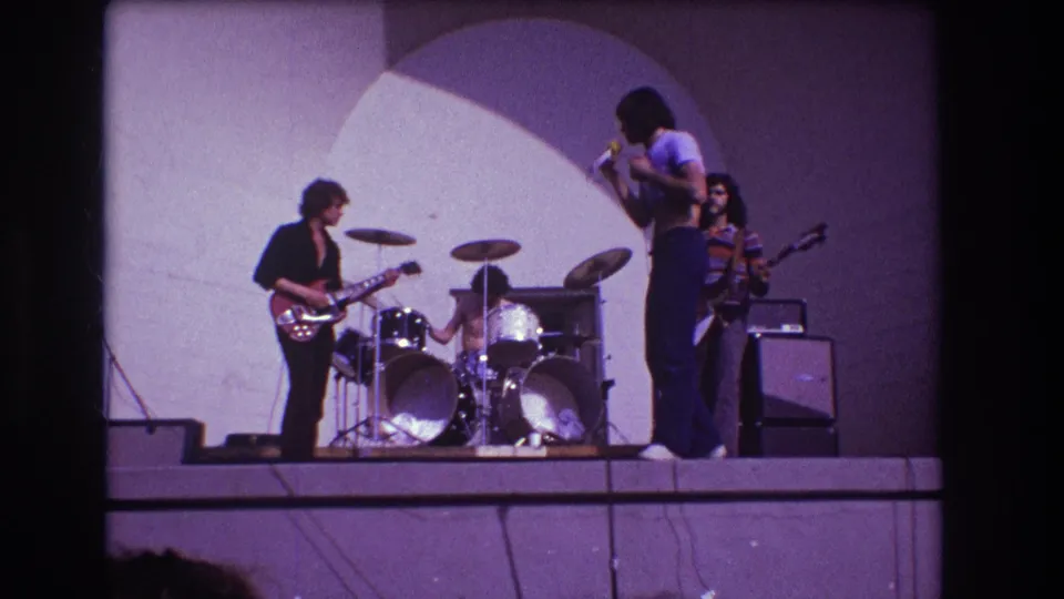 1973 Medford, New Jersey 8mm frame showing a Black male singer with microphone beside a guitarist, drummer, and bassist on an outdoor stage.