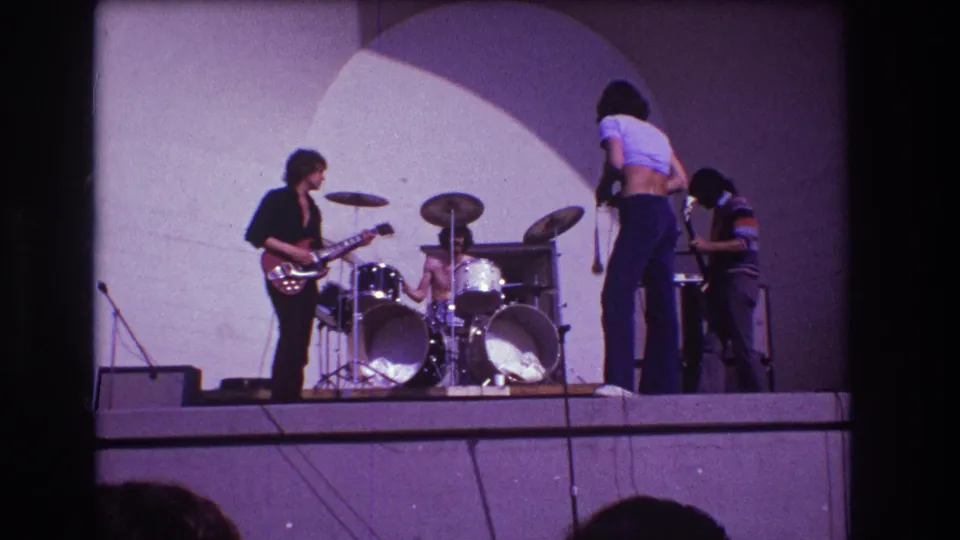 1973 Medford, New Jersey 8mm frame showing the singer near center stage, with guitarist, drummer, bassist, and amplifier arranged across the platform.