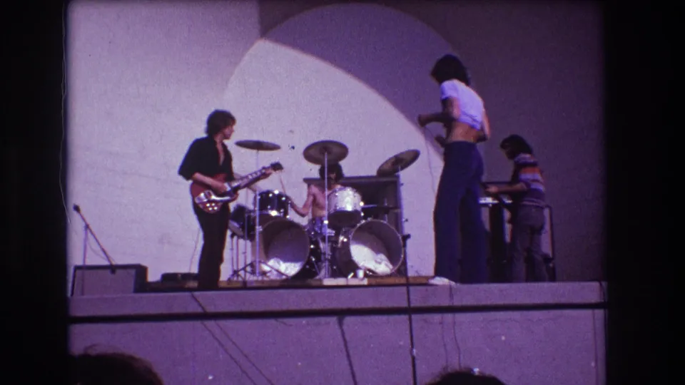1973 Medford, New Jersey frame of a live band on an outdoor stage, with the singer turned toward the bassist beside the amplifier.
