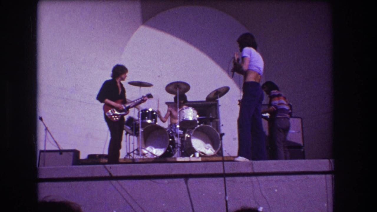 1973 Medford, New Jersey 8mm frame showing a small rock band on stage with guitarist at left, drummer at center, vocalist near a microphone, and keyboardist at right.