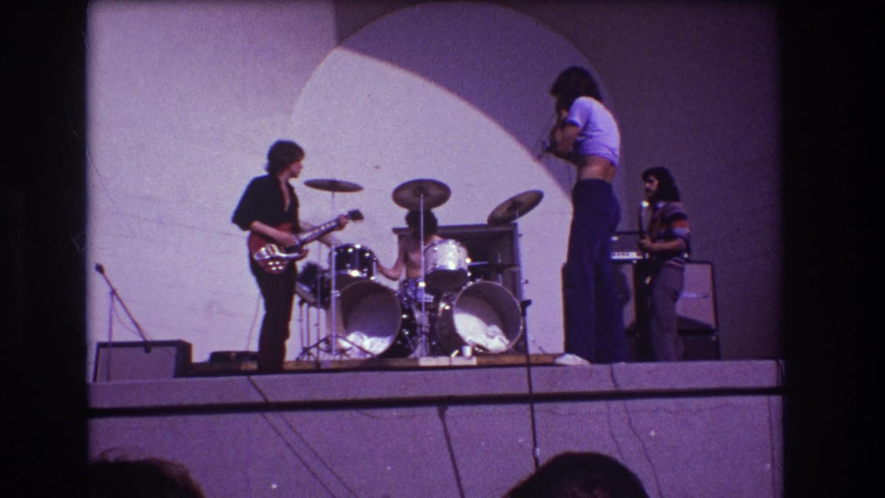 Four musicians stand across a stage with drums, guitar, keyboard, microphone stands, and amplifiers.