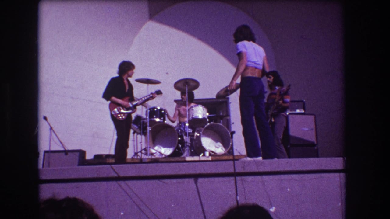Band members continue performing as the vocalist turns near the drum kit and the guitarist plays at left.