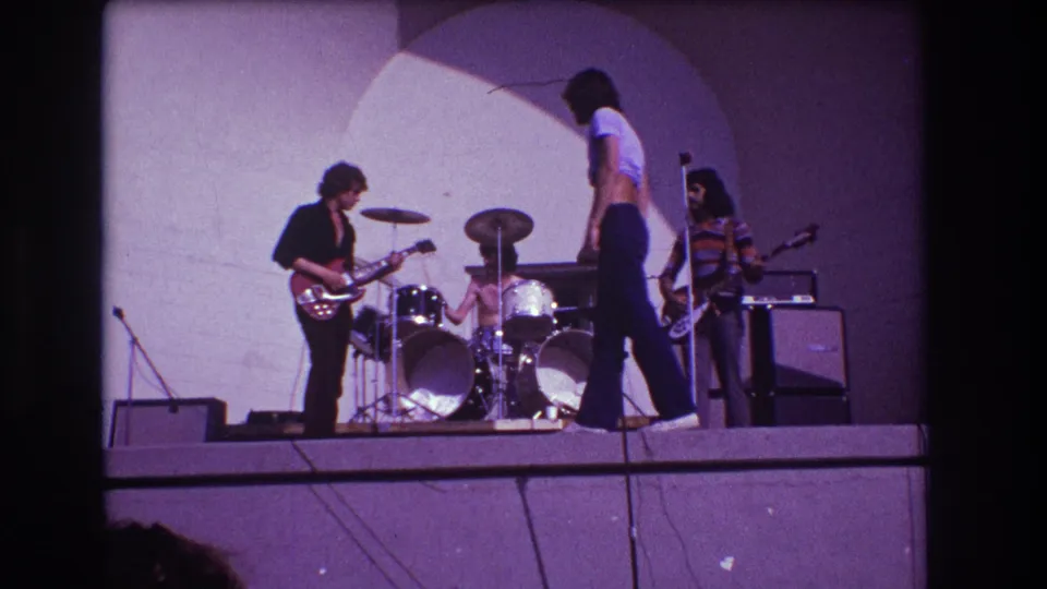 1973 Medford, New Jersey frame of a small rock band, with the vocalist walking across the stage in front of the drummer while guitarist and keyboardist remain at their instruments.