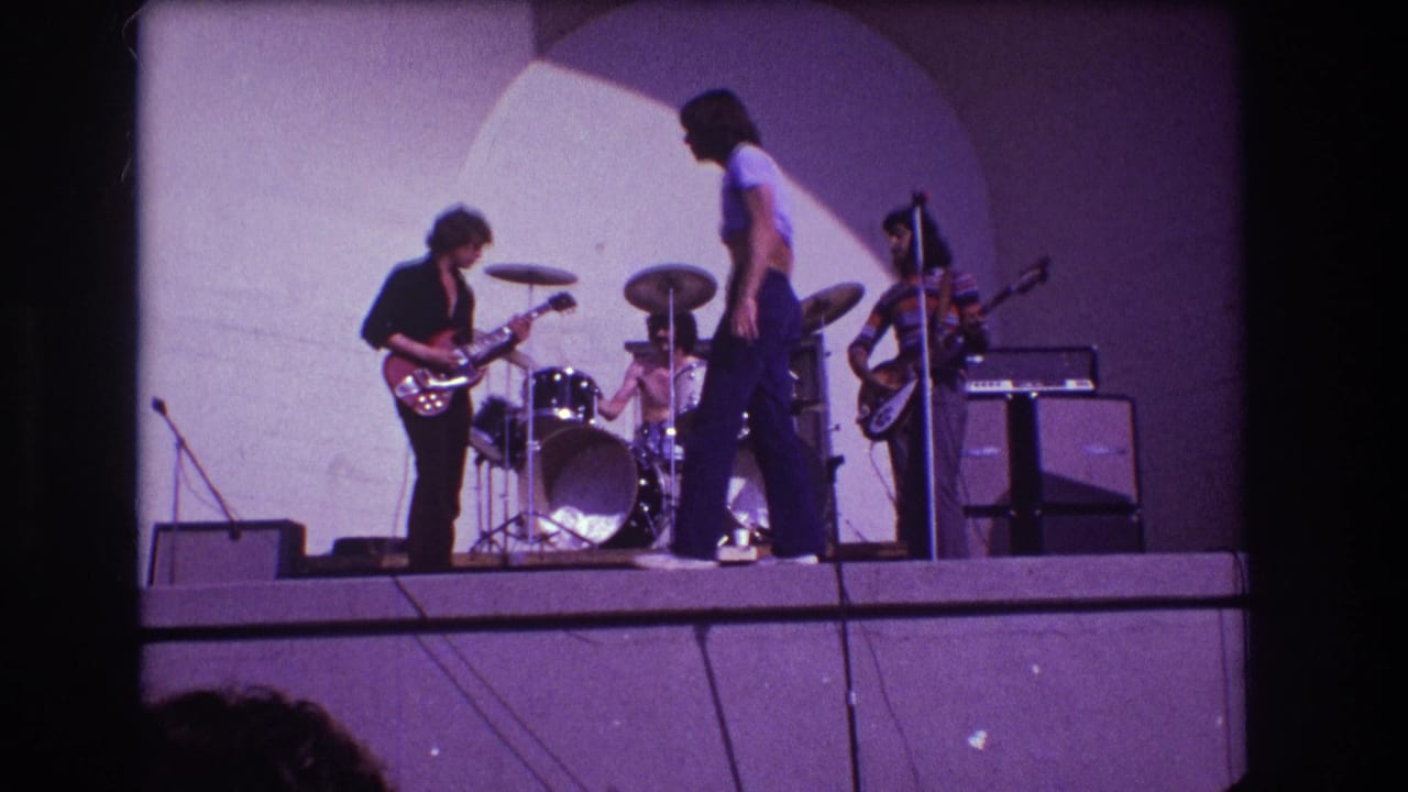 Rock band on a stage with guitarist, drummer, vocalist, and another musician under an arched backdrop.