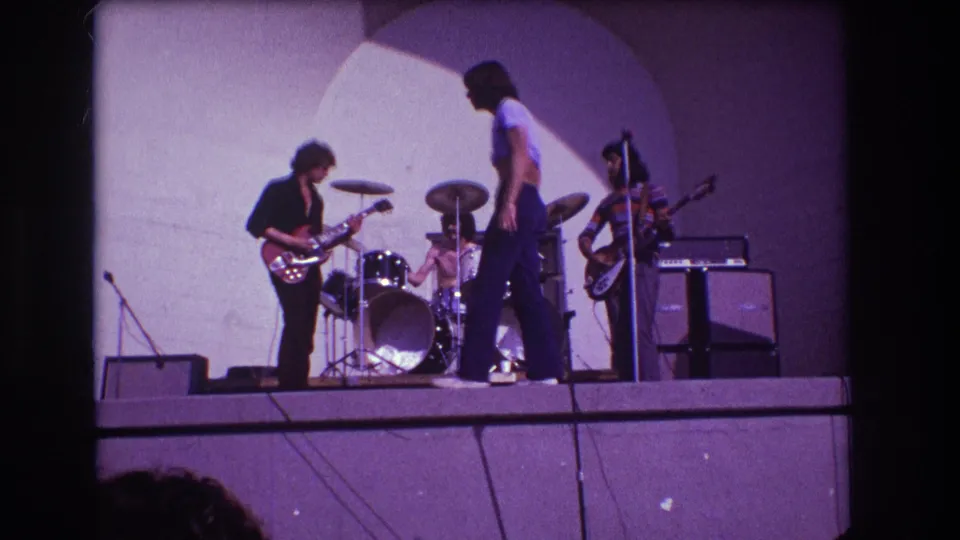1973 Medford, New Jersey 8mm frame showing a small rock band performing on stage with guitar, drums, vocalist, amplifiers, and cables.