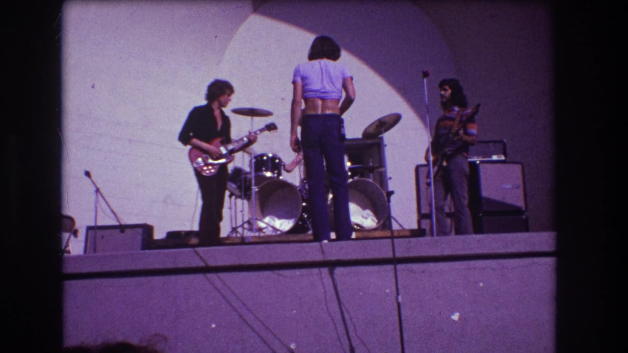 Rock band on a stage with guitarist, drummer, bassist, amplifiers, and a spotlighted wall.
