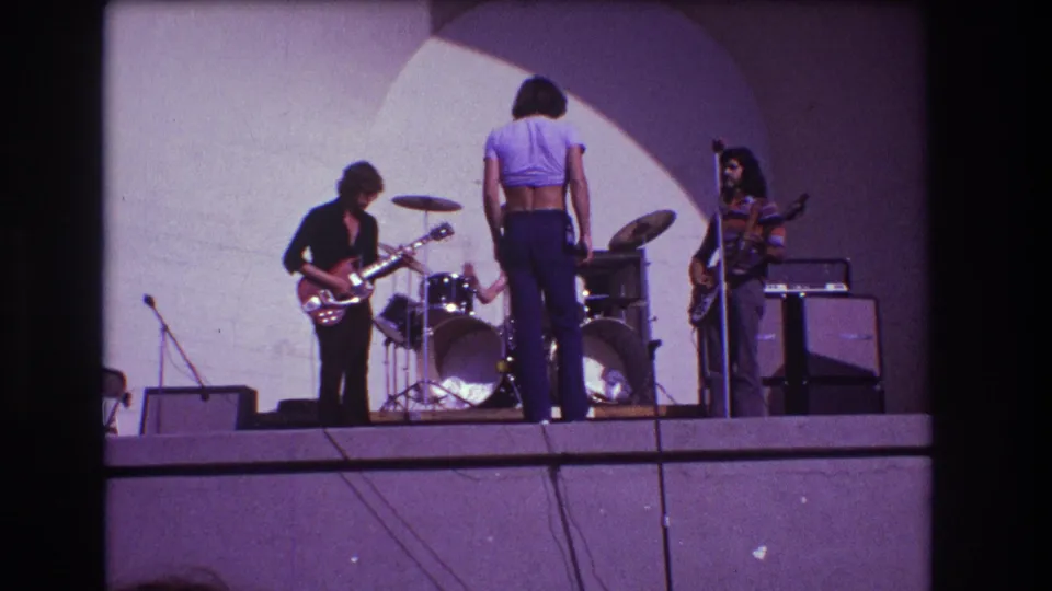 Home-movie still from the 1973 Medford performance showing the trio on stage with visible grain, soft focus, and purple tint.