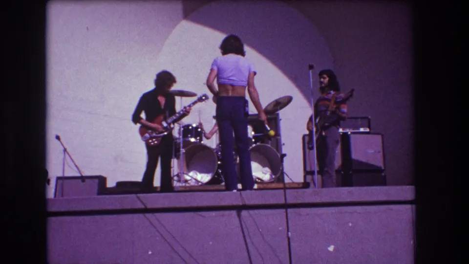 1973 Medford, New Jersey frame of a small rock band performing on a plain stage, seen through dark 8mm film borders.
