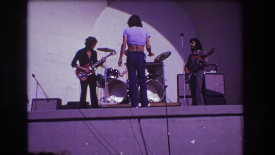 1973 Medford, New Jersey 8mm frame showing a small rock band rehearsing on stage with guitars, drums, microphones, and amplifiers.