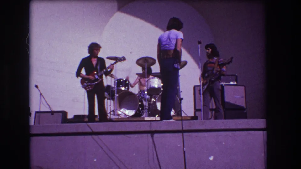 1973 Medford, New Jersey frame showing the drummer more clearly between guitarists as the band rehearses on a raised stage.