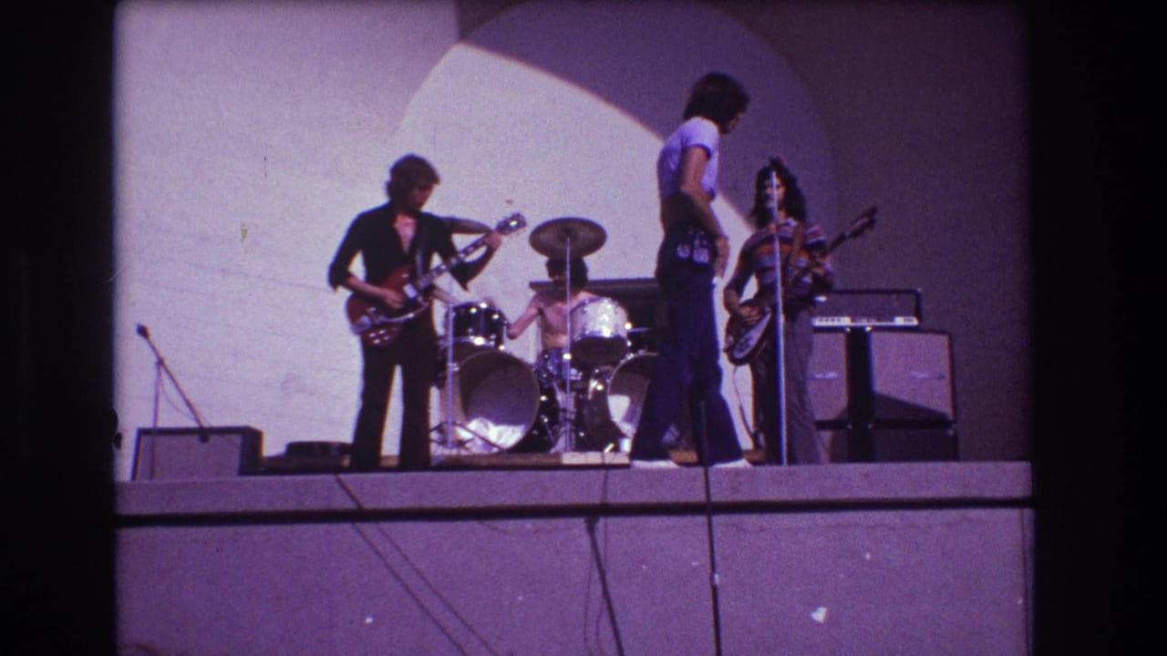 1973 Medford, New Jersey 8mm frame showing a rock band performing on a simple outdoor stage beneath a pale arch backdrop.