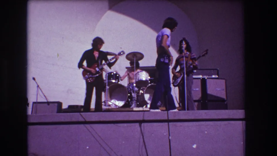 1973 Medford, New Jersey 8mm frame showing a rock band performing on a simple outdoor stage beneath a pale arch backdrop.