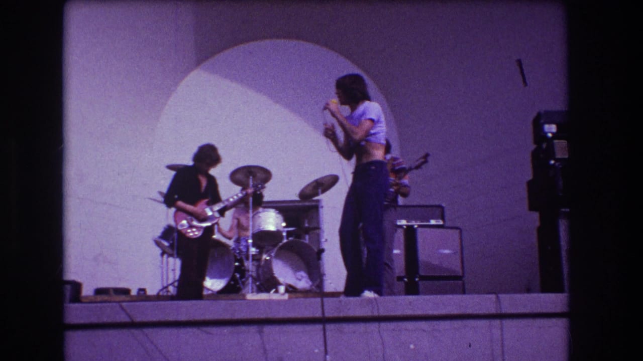 1973 Medford, New Jersey 8mm frame showing a rock singer at a microphone beside guitarist, drummer, amps, and a circular stage backdrop.