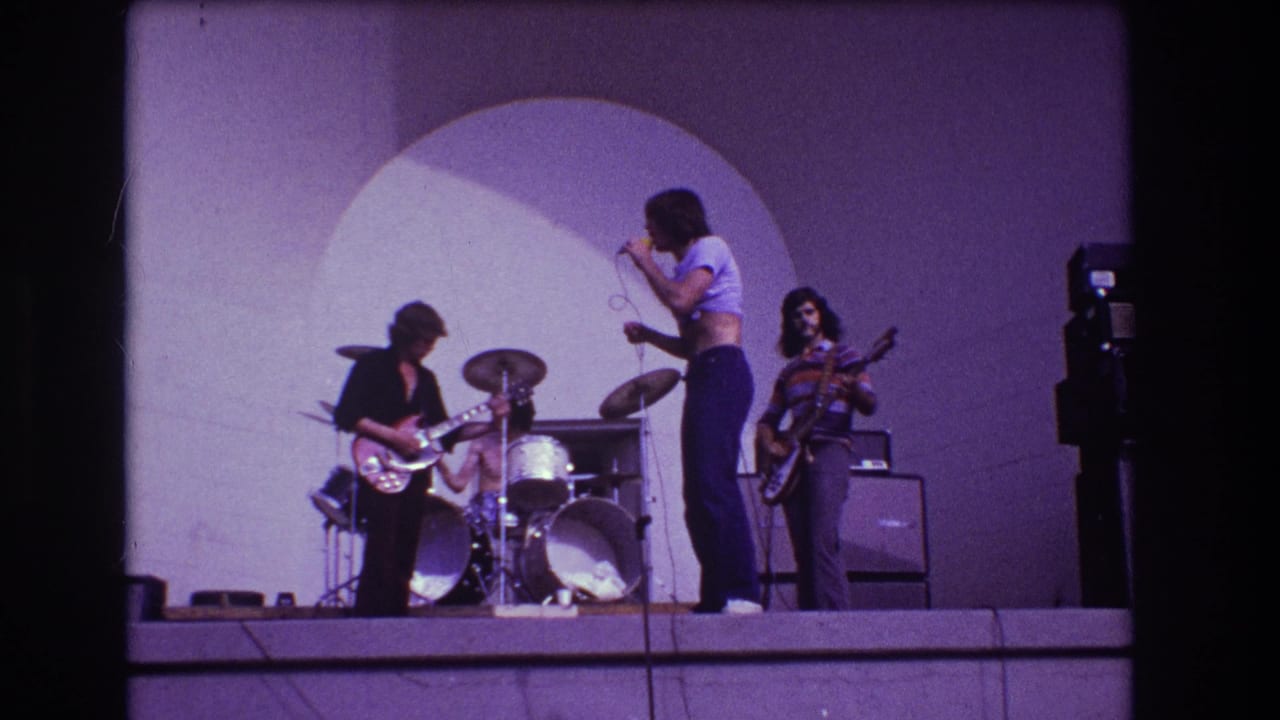 1973 Medford, New Jersey frame with singer holding a microphone as guitarist, drummer, and bassist play on a purple-lit stage.