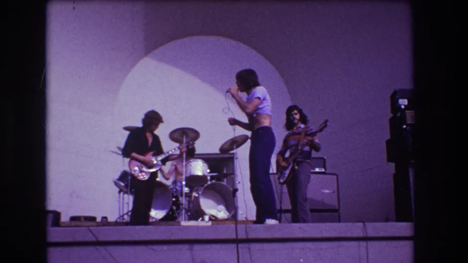 1973 Medford, New Jersey frame with singer holding a microphone as guitarist, drummer, and bassist play on a purple-lit stage.