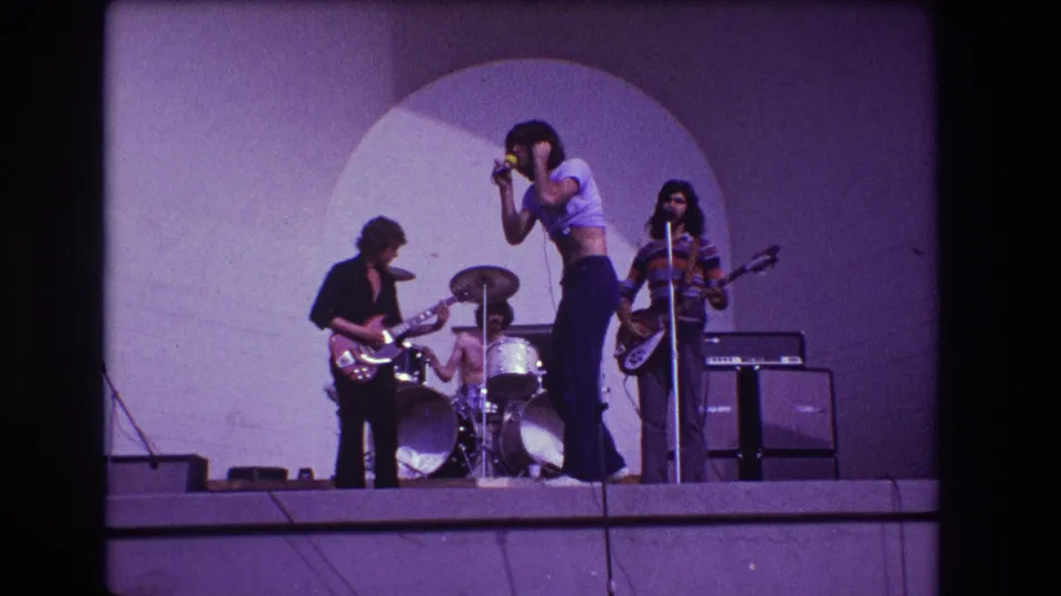Closer 8mm view of the Medford outdoor concert, with the singer in flared pants framed between guitar and bass players.