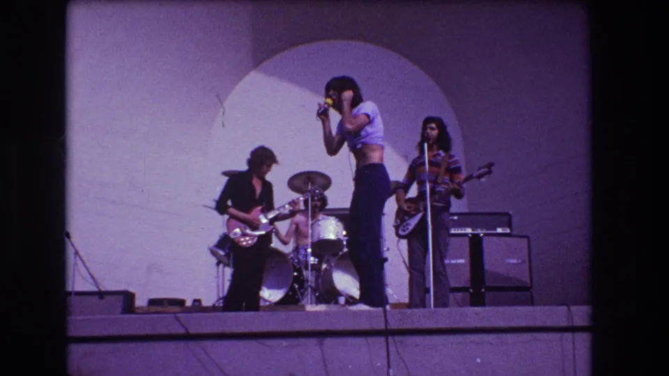 Purple-tinted 8mm frame of the 1973 Medford band performance with vocalist, guitarist, bassist and drummer visible.