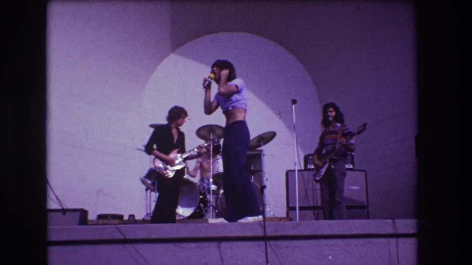 1973 Medford, New Jersey stage frame showing the singer, guitarist, drummer, and bassist spread across a purple-toned rock performance setup.