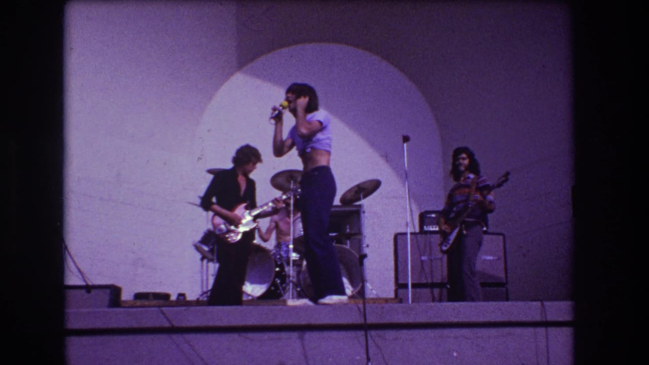 Rock band performing on an outdoor stage with singer, guitarist, drummer, and amplifiers visible.