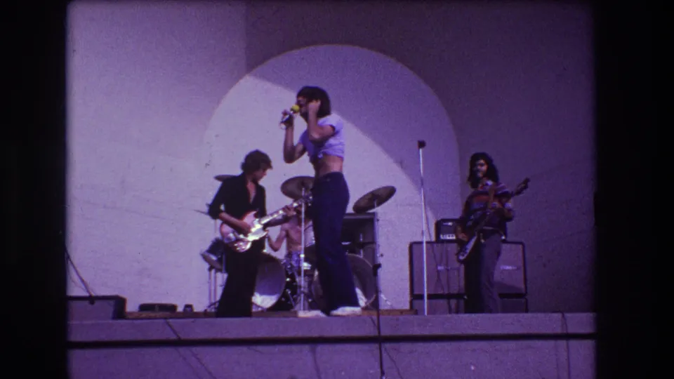 1973 Medford, New Jersey home-movie frame of a rock band on an outdoor stage, with the singer at a microphone and guitarists beside a drum kit.
