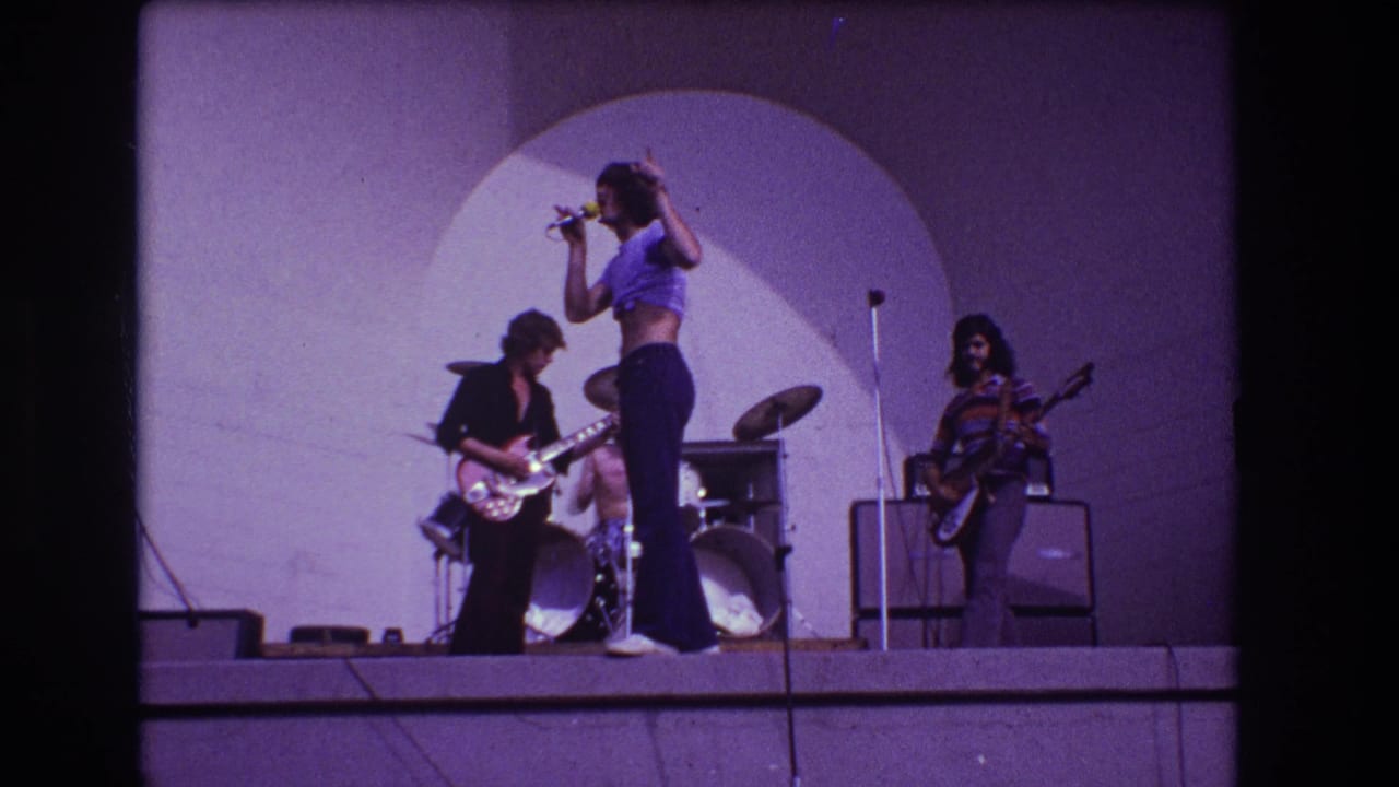 1973 Medford, New Jersey frame showing the singer gesturing during an outdoor rock performance with guitarists, drums, and amplifiers visible.
