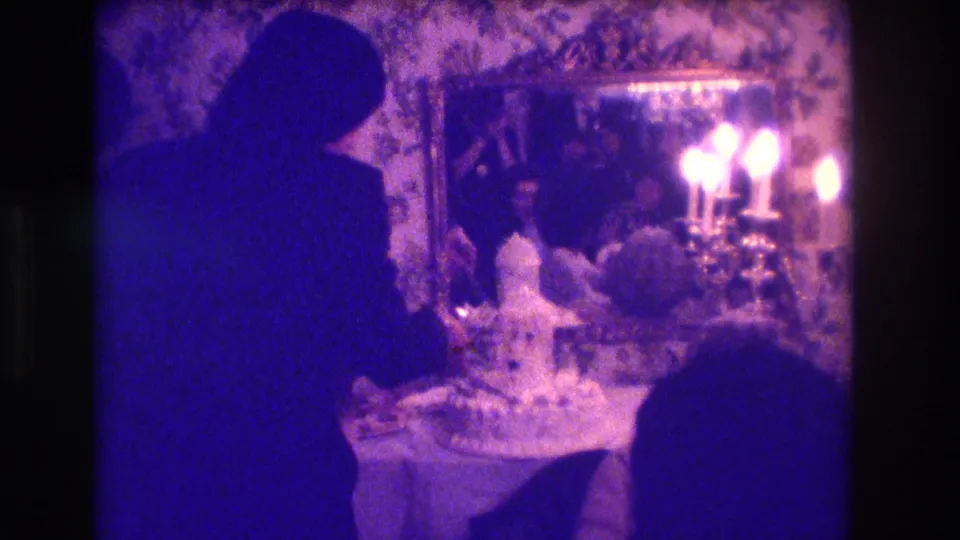 The couple leans over the wedding cake in a grainy 1973 Medford home-movie frame with purple-blue color cast and mirrored candles.