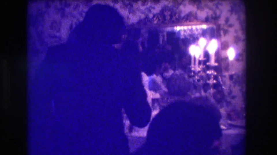1973 Medford, New Jersey home-movie frame of silhouetted wedding participants around a candlelit cake table in a floral-wallpapered interior.