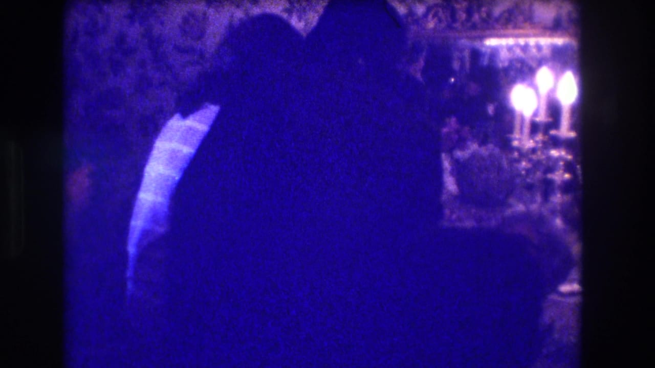 In the supplied 1973 Medford home-movie clip, silhouetted adults gather in a dim formal interior beside patterned wallpaper and candelabra light.