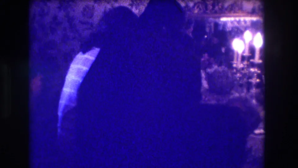 In the supplied 1973 Medford home-movie clip, silhouetted adults gather in a dim formal interior beside patterned wallpaper and candelabra light.