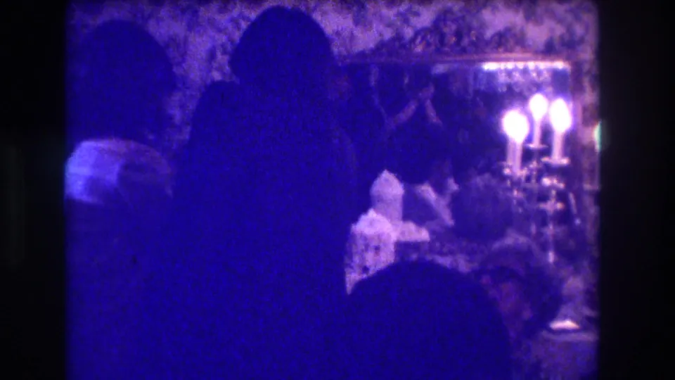 Several people cluster around an older woman in the supplied 1973 Medford home-movie frame, with candelabra light and ornate wallpaper visible.