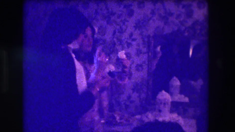1973 Medford home-movie frame showing dark human silhouettes around a table with a decorated cake-like object in a private interior.