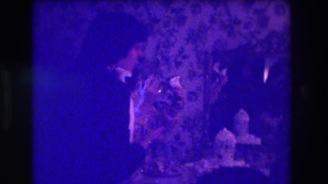 1973 Medford home-movie frame showing adults in a dark room near candles and floral wallpaper, with heavy purple color cast.