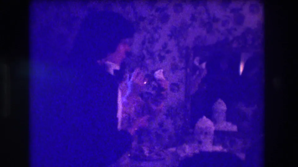 1973 Medford home-movie frame showing adults in a dark room near candles and floral wallpaper, with heavy purple color cast.