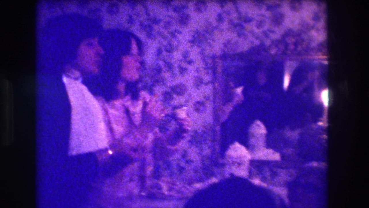 1973 Medford 8mm frame of a woman in a light dress and a man in a dark suit near a candlelit table.