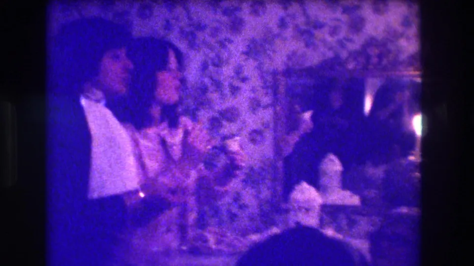 1973 Medford 8mm frame of a woman in a light dress and a man in a dark suit near a candlelit table.