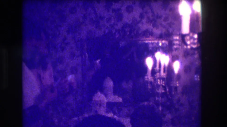 Low-light 1973 Medford home-movie frame with a bright candelabra, table setting, and partial adult figures in a private room.